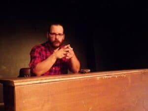 white man sitting at a desk on a theater stage